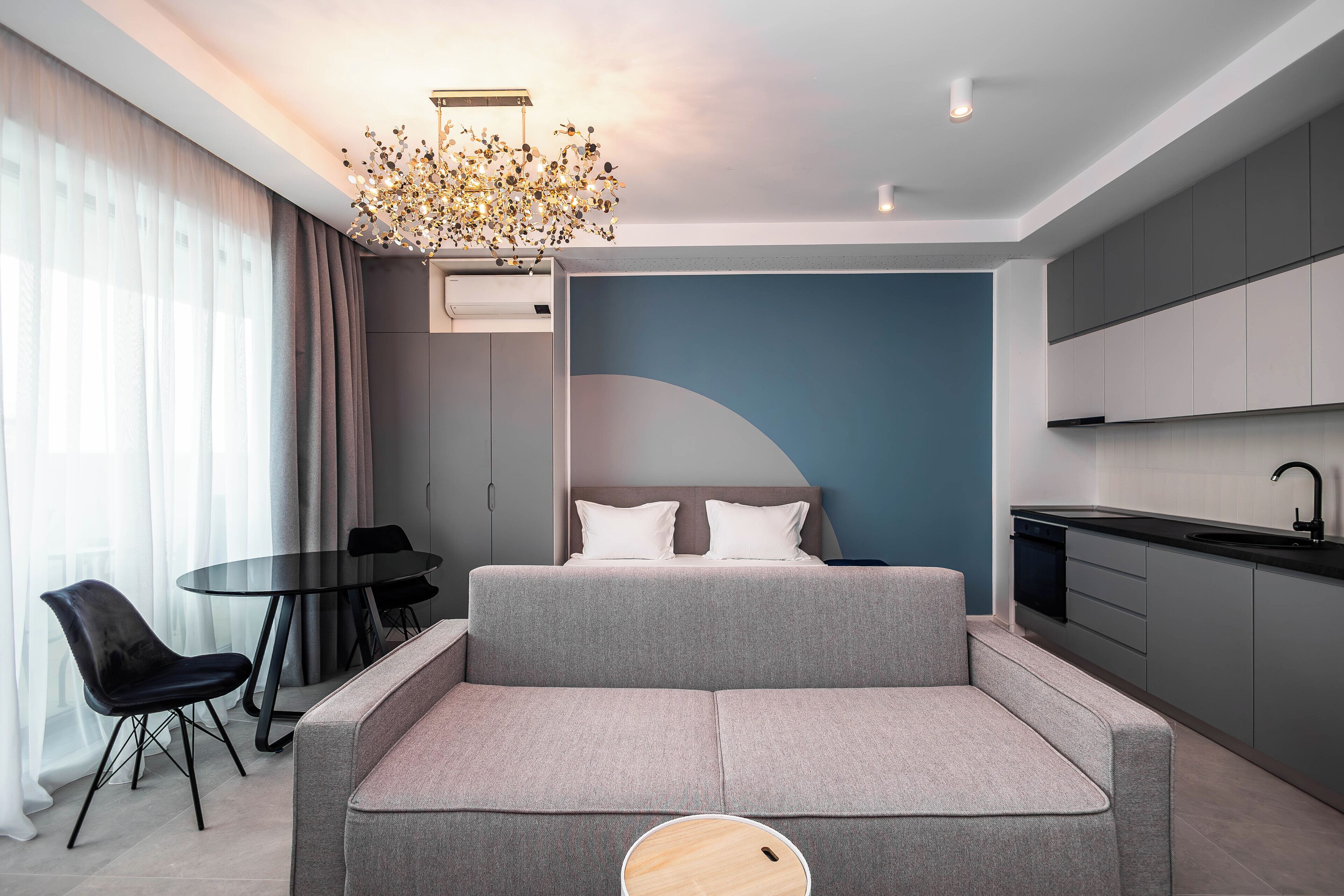 A modern bedroom with a sofa bed, a dining table with chairs, a kitchen area, and a large window with curtains.