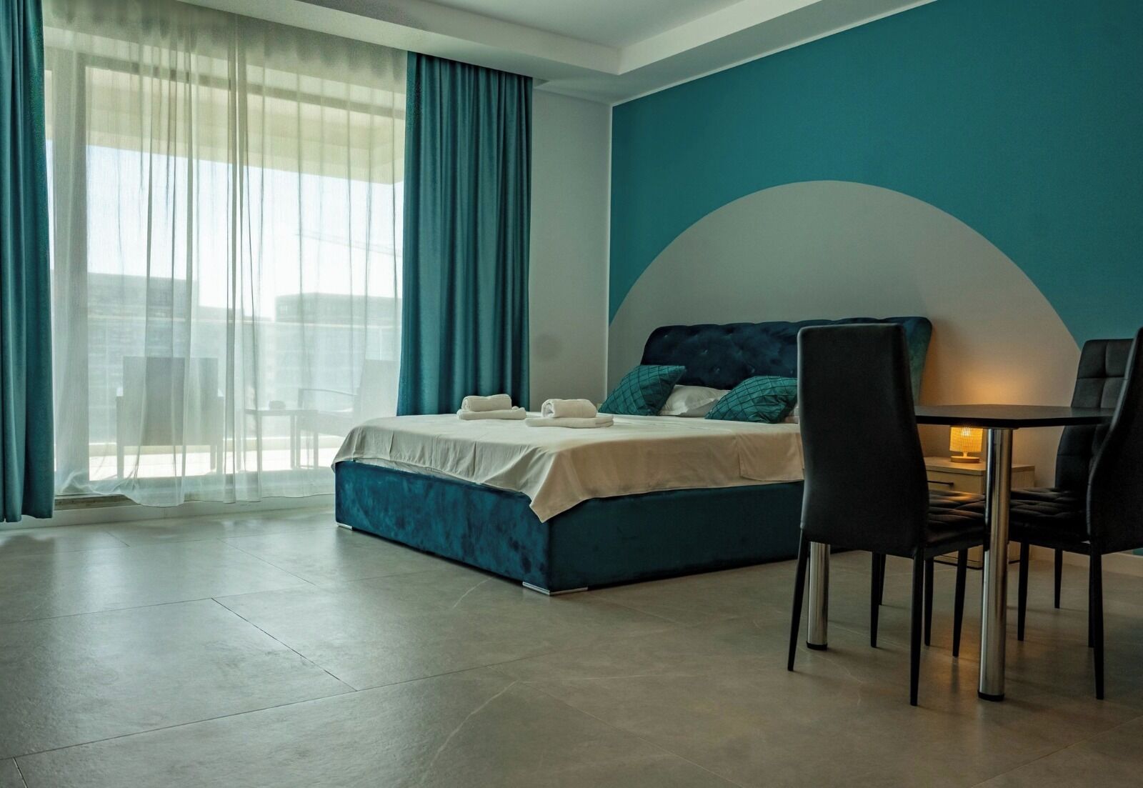 Habitació d'hotel moderna amb llit, taula de dinar amb cadires i vista a través d'una finestra amb cortines lleugeres.