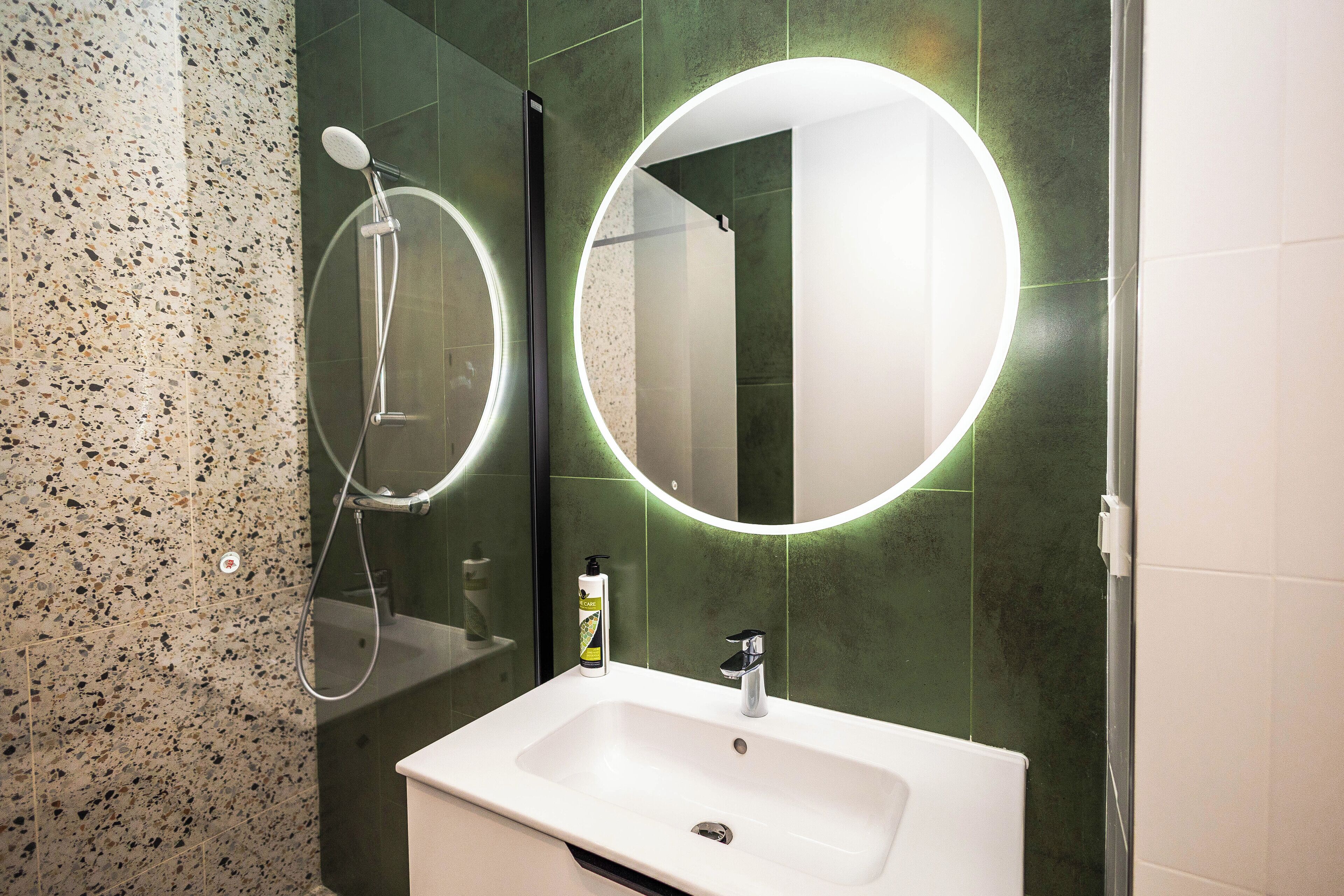 A bathroom with a white sink, a mirror with a circular light, and a shower area.