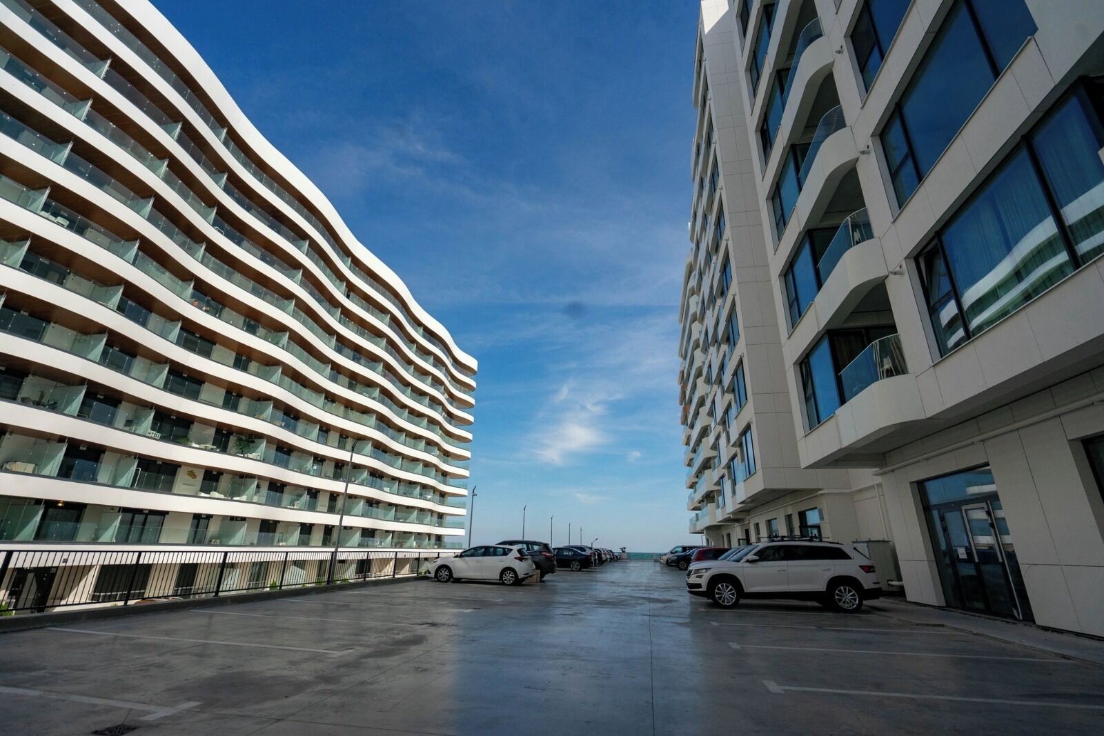 Un bâtiment moderne à la façade incurvée, doté de nombreux balcons et d’un espace de stationnement où sont garées des voitures.