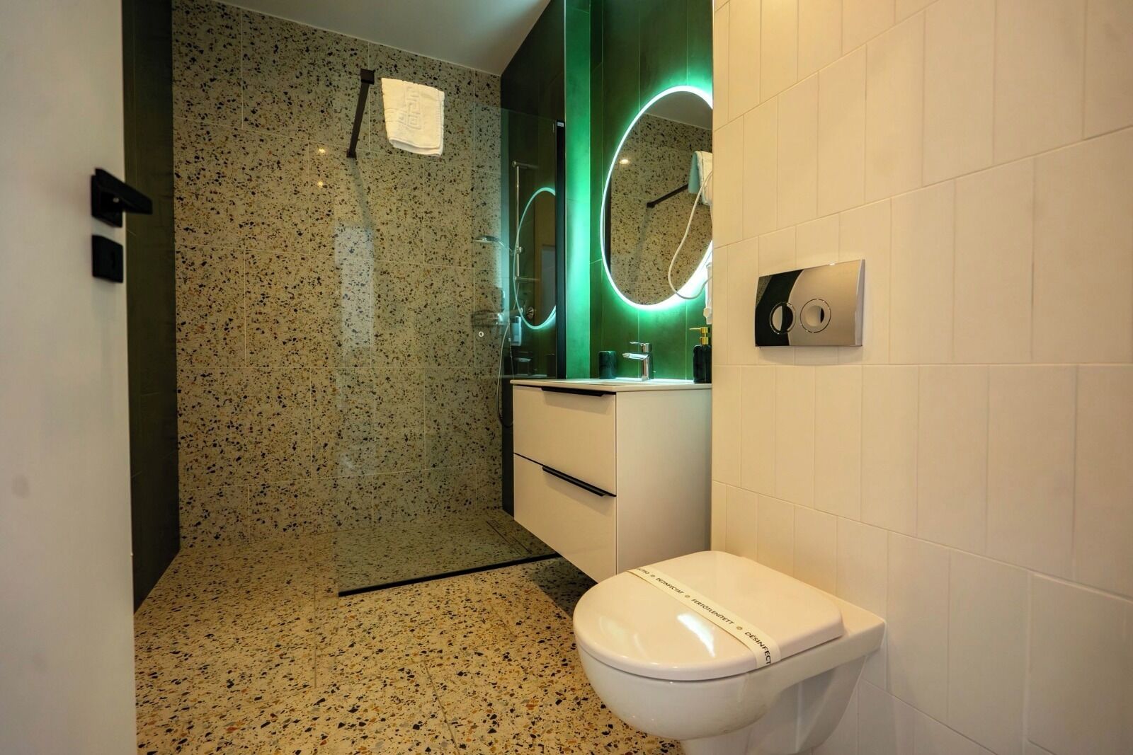 A modern bathroom with a granite speckled floor, a white toilet, a vanity with a mirror, and a green accent wall.