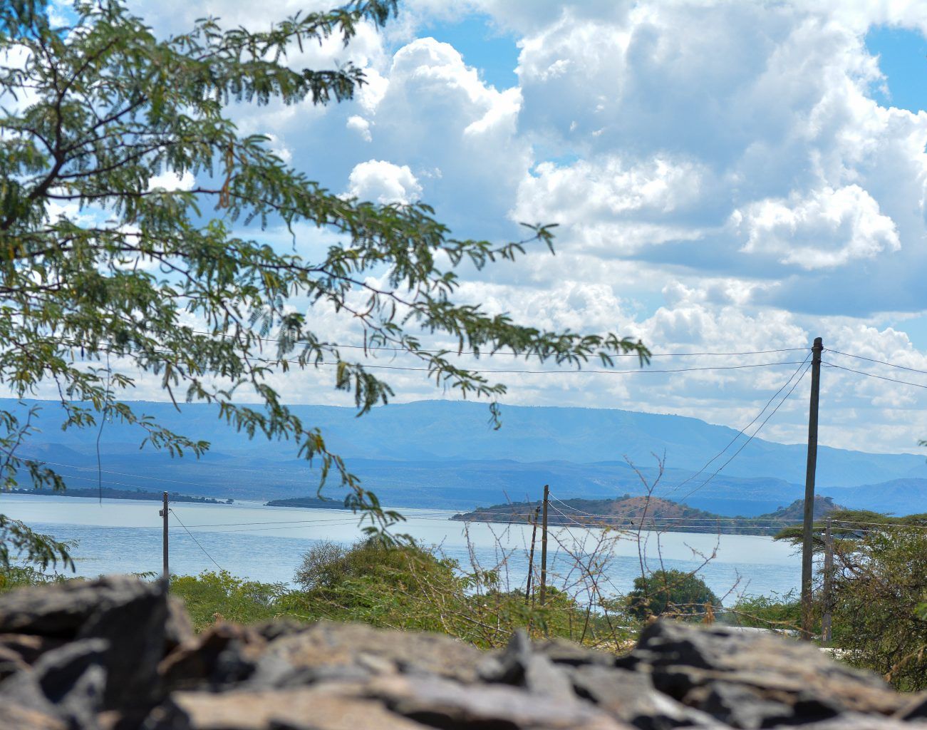 A scenic view of a lake with mountains in the background, power lines, and trees in the foreground.