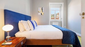 Un dormitorio con cama, mesita de noche, lámpara y ventana con vistas a edificios.
