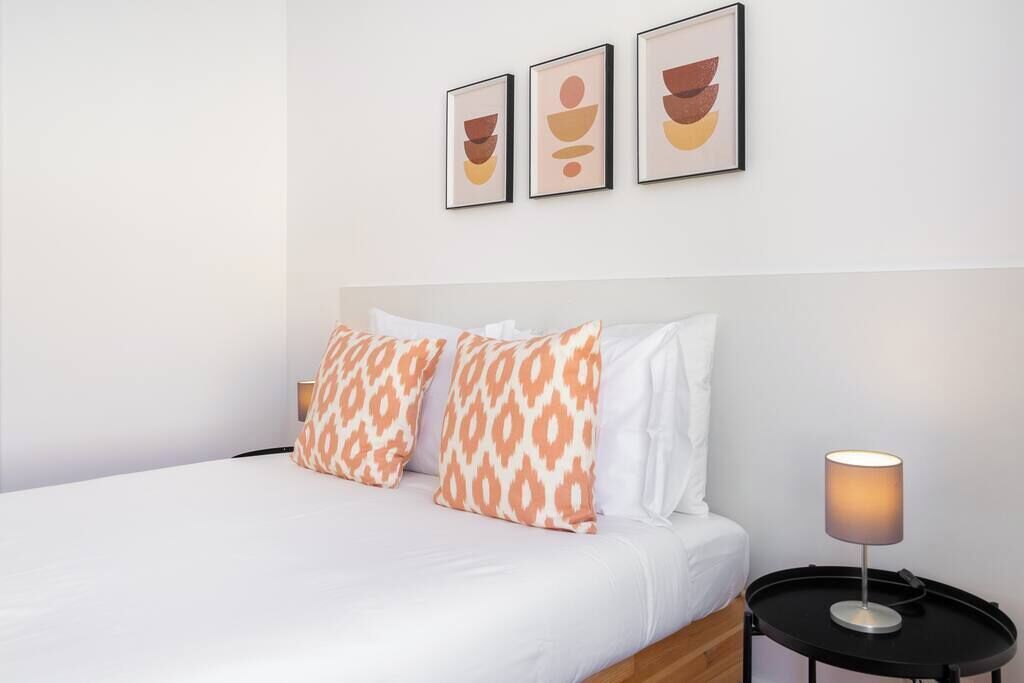 Una cama bien tendida con almohadas estampadas, una mesita de noche con lámpara y tres obras de arte abstracto sobre la cama.