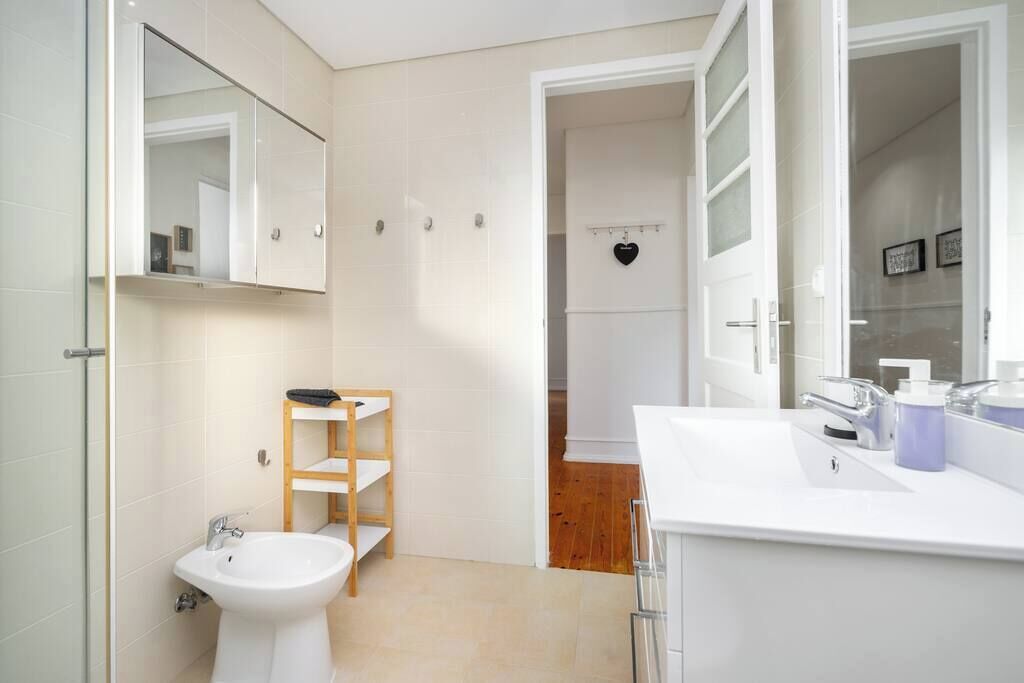 A modern bathroom with a white sink, a bidet, and a mirror. There is a wooden shelf unit and a heart-shaped decoration on the wall.
