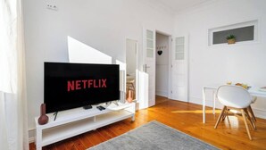 Un soggiorno moderno con un'ampia televisione a schermo piatto che mostra 'NETFLIX', un mobile TV bianco, un'area pranzo con tavolo e sedia bianchi e un corridoio che conduce a una stanza con una decorazione a forma di cuore.