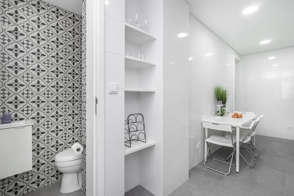 A bathroom with a toilet, a sink, and a shower area. There is a white table with chairs and a potted plant. Shelves are mounted on the wall.