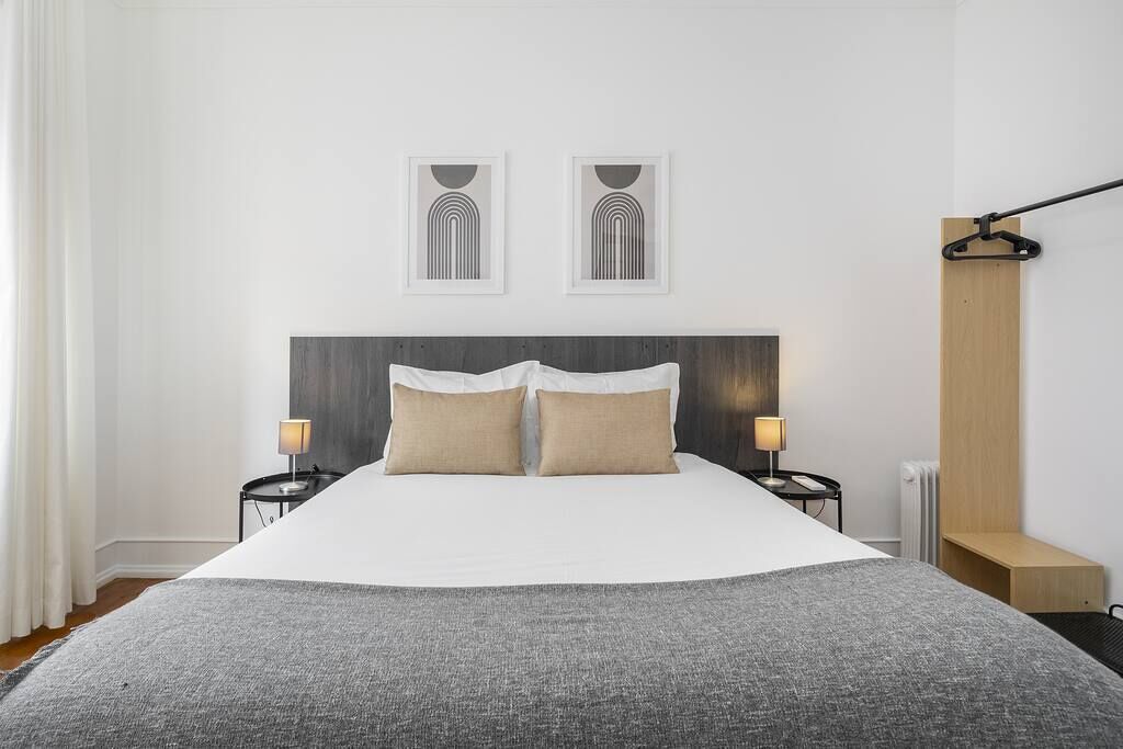 Een netjes opgemaakt bed met een grijze deken, twee nachtkastjes met lampen, en twee ingelijste kunstwerken erboven.