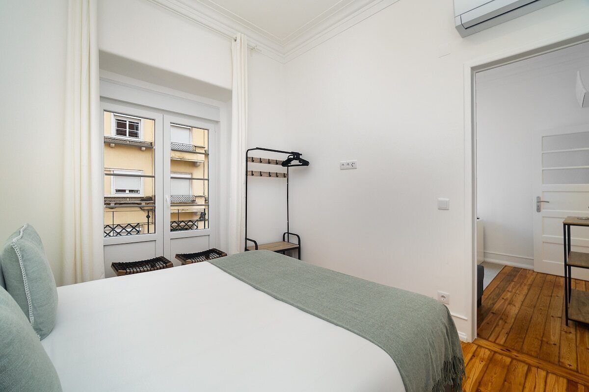 Un dormitorio con cama, radiador, ventana con vista a un edificio y puerta que da a otra habitación.