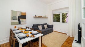 Un salon moderne comprenant un coin repas, un canapé gris, un téléviseur et une grande fenêtre avec des rideaux.