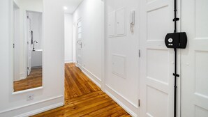 Un couloir aux murs blancs et au plancher en bois, qui mène à une salle de bain équipée d’une douche et d’un lavabo.