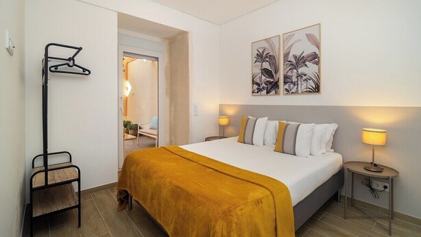A bedroom with a bed, a bedside table with a lamp, a coat rack, and a door leading to another room.