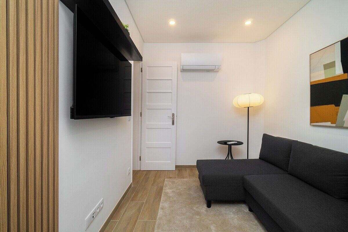 Un salon moderne avec un canapé d’angle, un téléviseur à écran plat fixé au mur, une lampe sur pied et un appareil de climatisation.