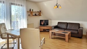 A modern living room with a sofa, a dining table, chairs, a TV, and a chandelier. - Baltique - Holiday apartment 3 Glowe - Holiday apartment Baltique 3 (Glowe)
