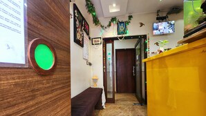 A reception area with a wooden desk, a bench, a wall calendar, and a television mounted on the wall. - Bhammars Inn - A Pure Veg Boutique Hotel (Kolkata)