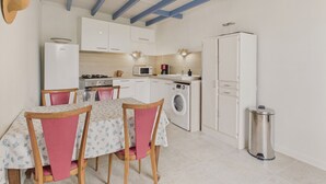 Une cuisine compacte avec des meubles blancs, un four à micro-ondes, une cuisinière et un lave-linge. On y trouve une table à manger avec des chaises, une poubelle et un plafond avec des poutres apparentes.