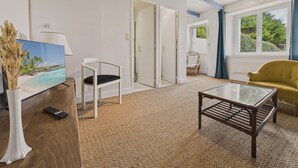 Une pièce avec une table basse en bois, une chaise blanche, un fauteuil jaune, un tableau représentant une plage et une porte donnant sur une autre pièce.