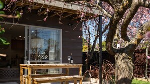 Um deck de madeira com mesa e banco, uma porta de vidro que dá para um quarto e uma árvore com flores cor-de-rosa.