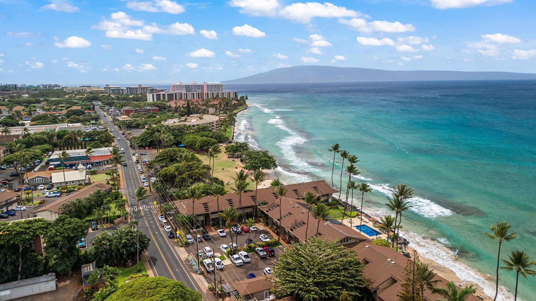 Un hotel frente al mar con vista a la playa, estacionamiento y una carretera con vehículos.