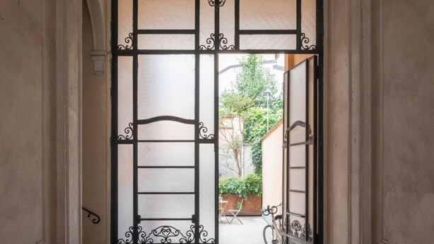Une porte surmontée d’un portail en métal décoratif, donnant sur un espace extérieur où se trouvent un vélo et de la verdure.
