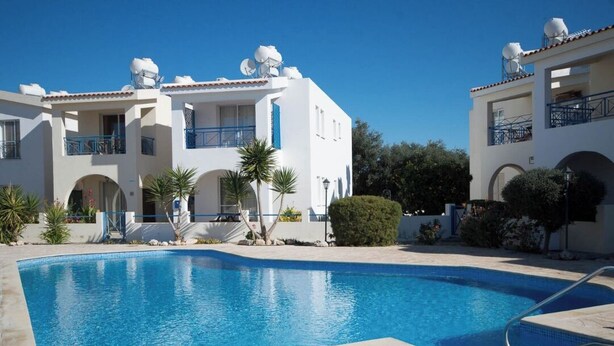 A swimming pool with a clear blue water, surrounded by a white building with balconies and satellite dishes on the roof.
