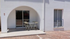 A building with a white exterior, a glass door entrance, a small patio with a table and chairs, and a window with a metal railing.