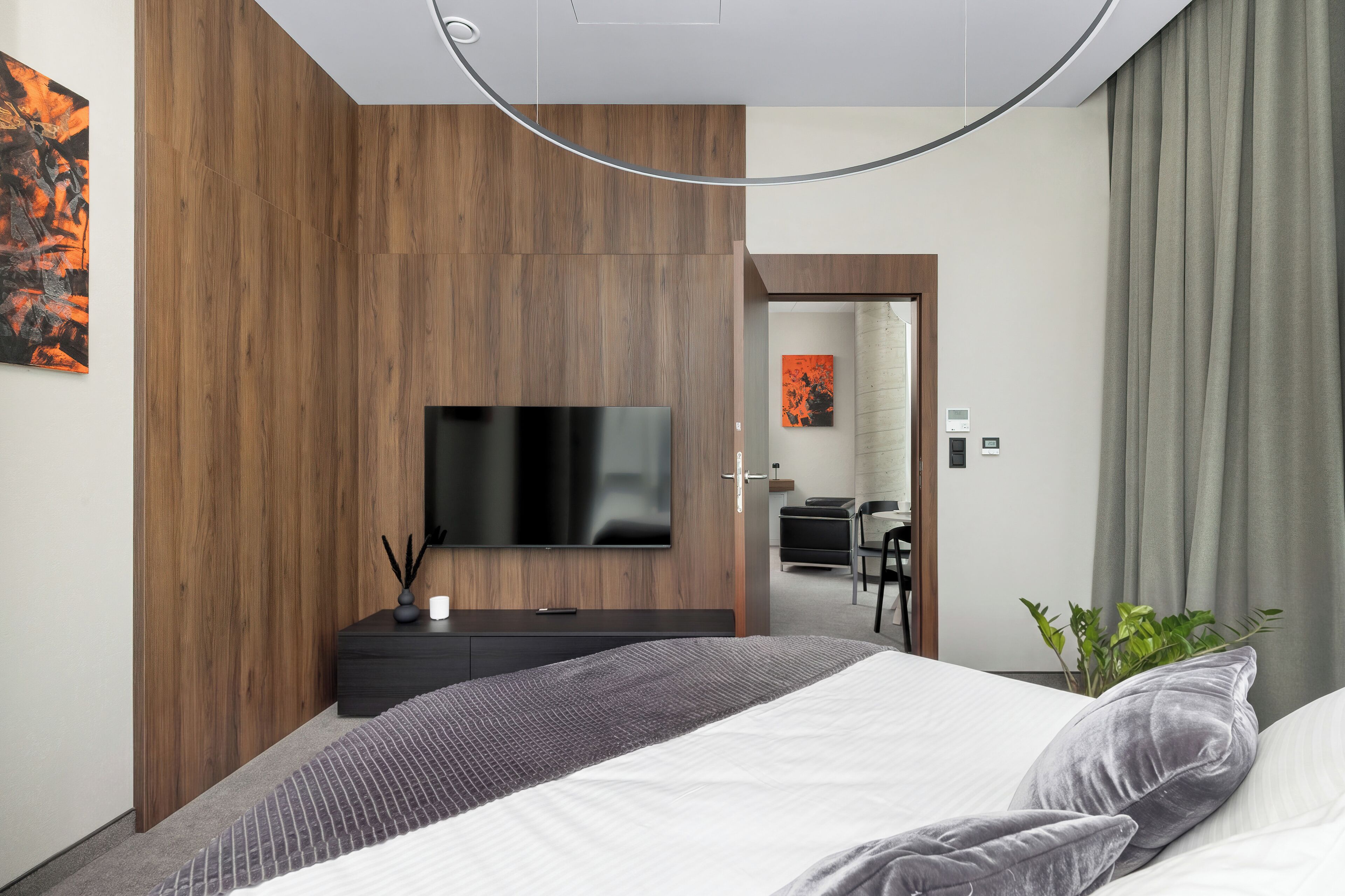 A modern hotel room with a large bed, a flat-screen TV mounted on a wooden wall, a small desk with a chair, and a plant.
