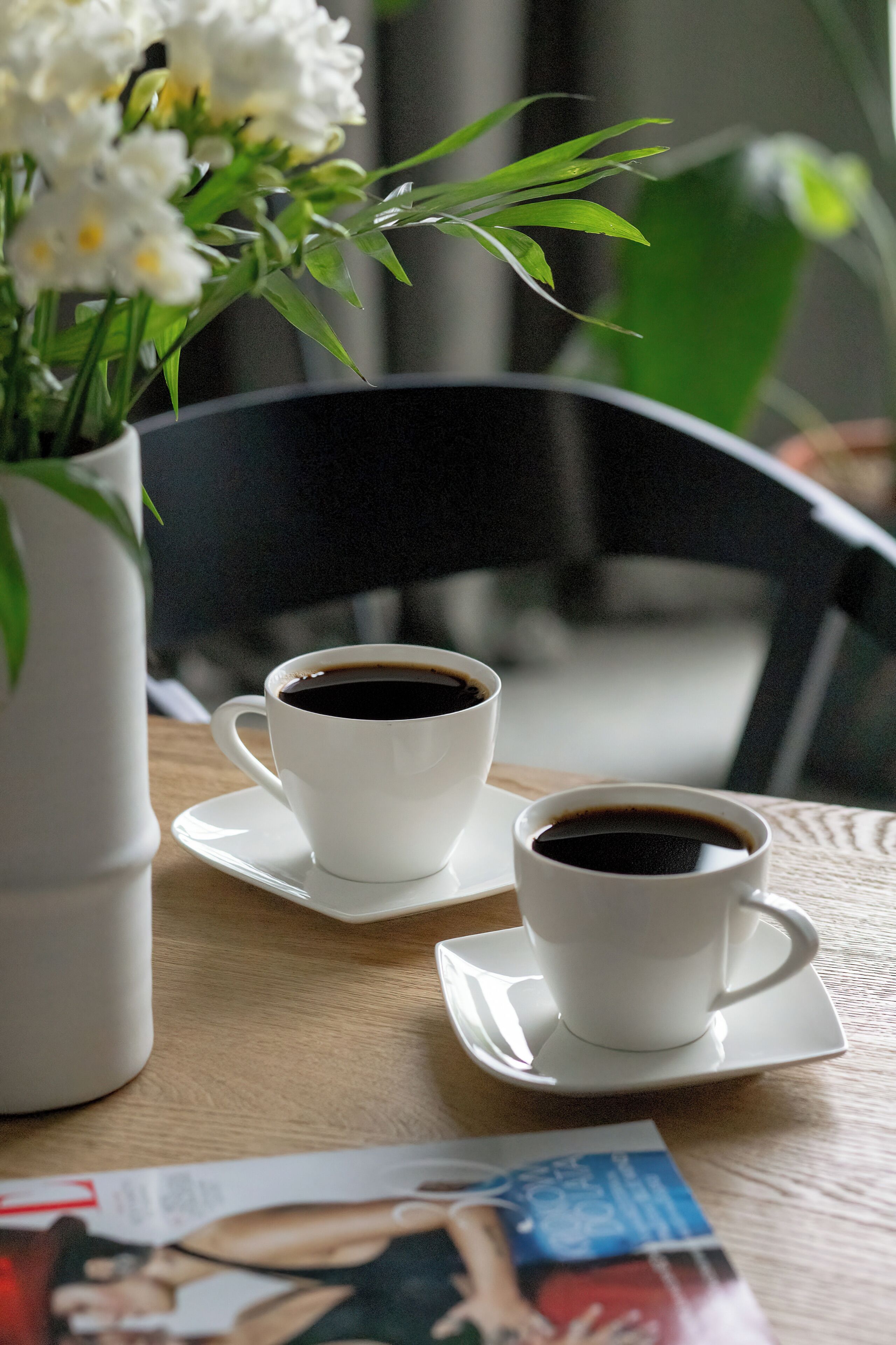 Two white cups of coffee on a table with a magazine, a white vase with flowers, and a black chair in the background.