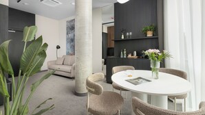A modern interior with a round dining table, cushioned chairs, a large plant, and a minimalist kitchen area.