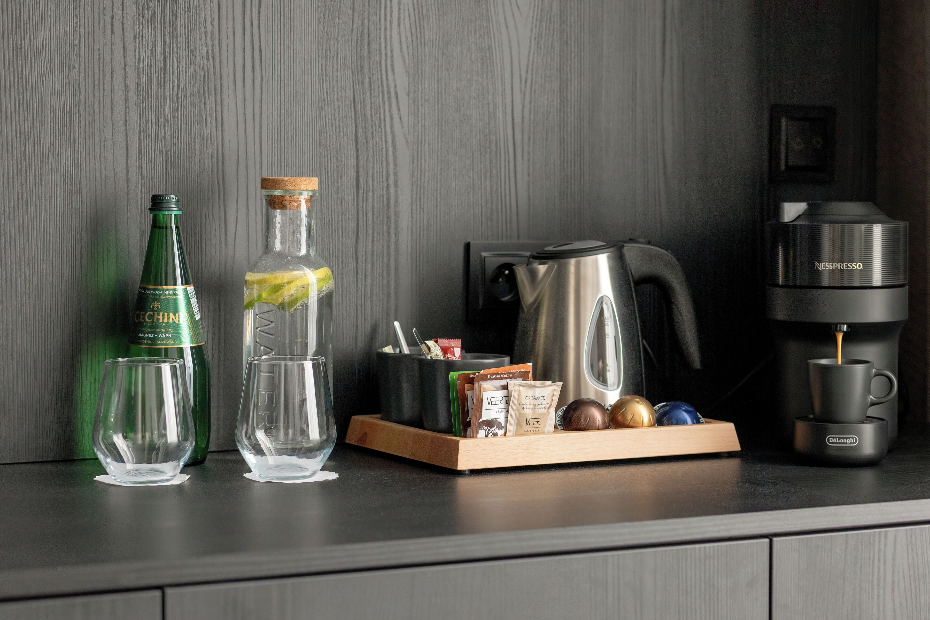 A kitchen counter with a coffee maker, a bottle of water, a glass, and a tray with coffee pods and a spoon.