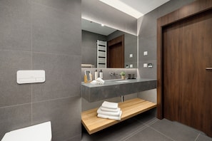 Un baño moderno con pared de azulejos grises, tocador de madera, lavamanos blanco, barra para toallas y banco de madera.