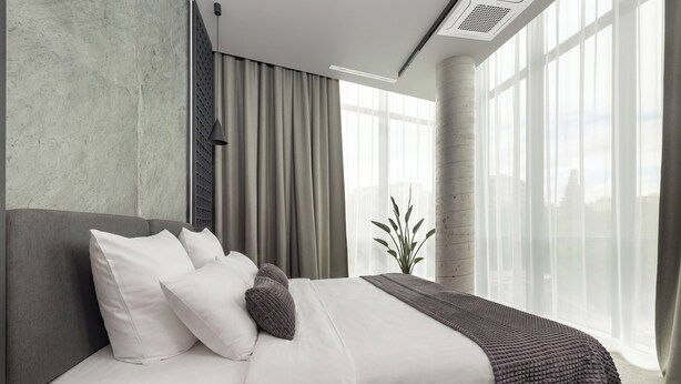 A modern hotel room with a large bed, a potted plant, and a view of the city through large windows.