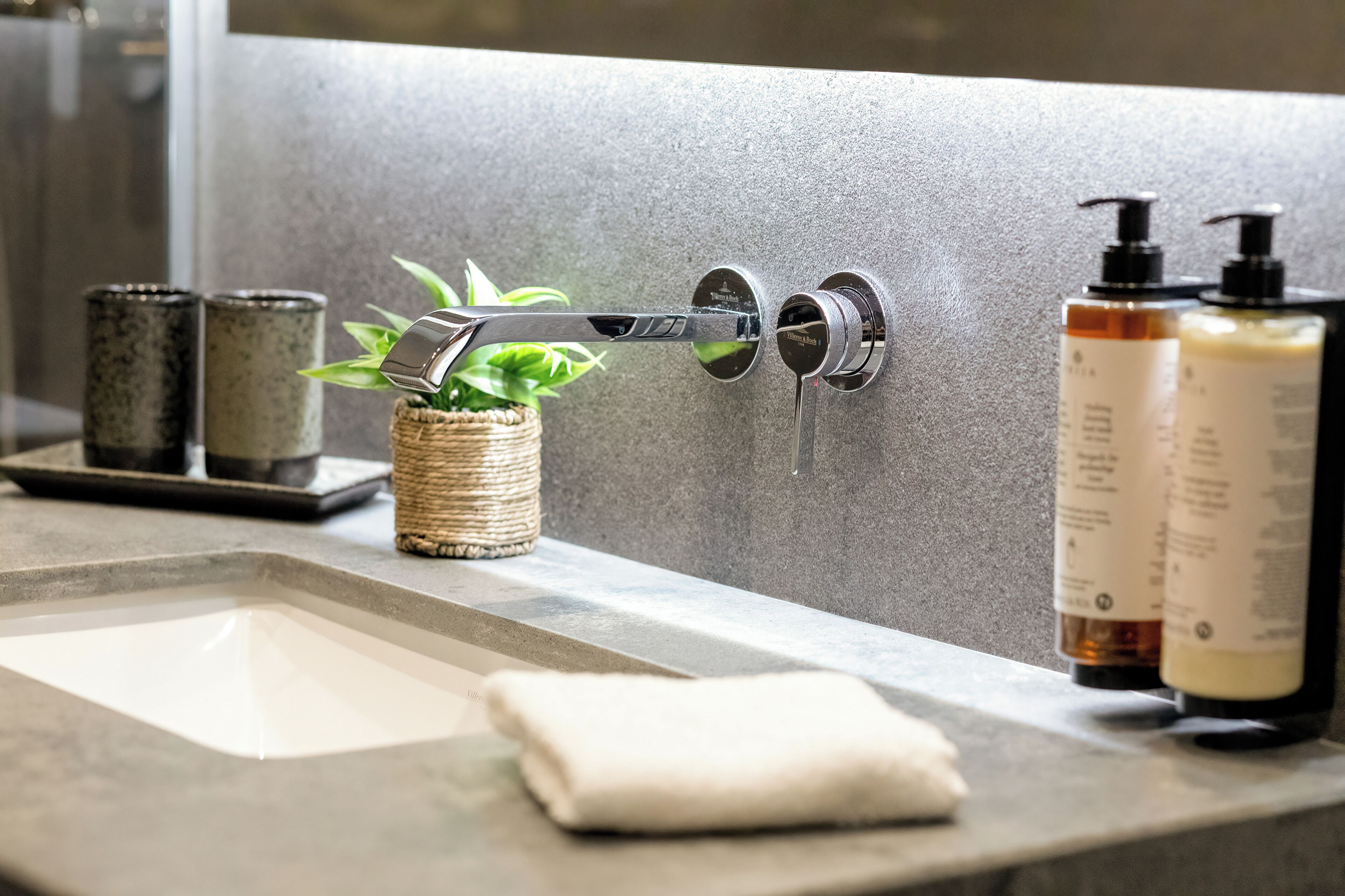 A bathroom with a modern faucet, a small plant, two soap dispensers, and a towel on the counter.