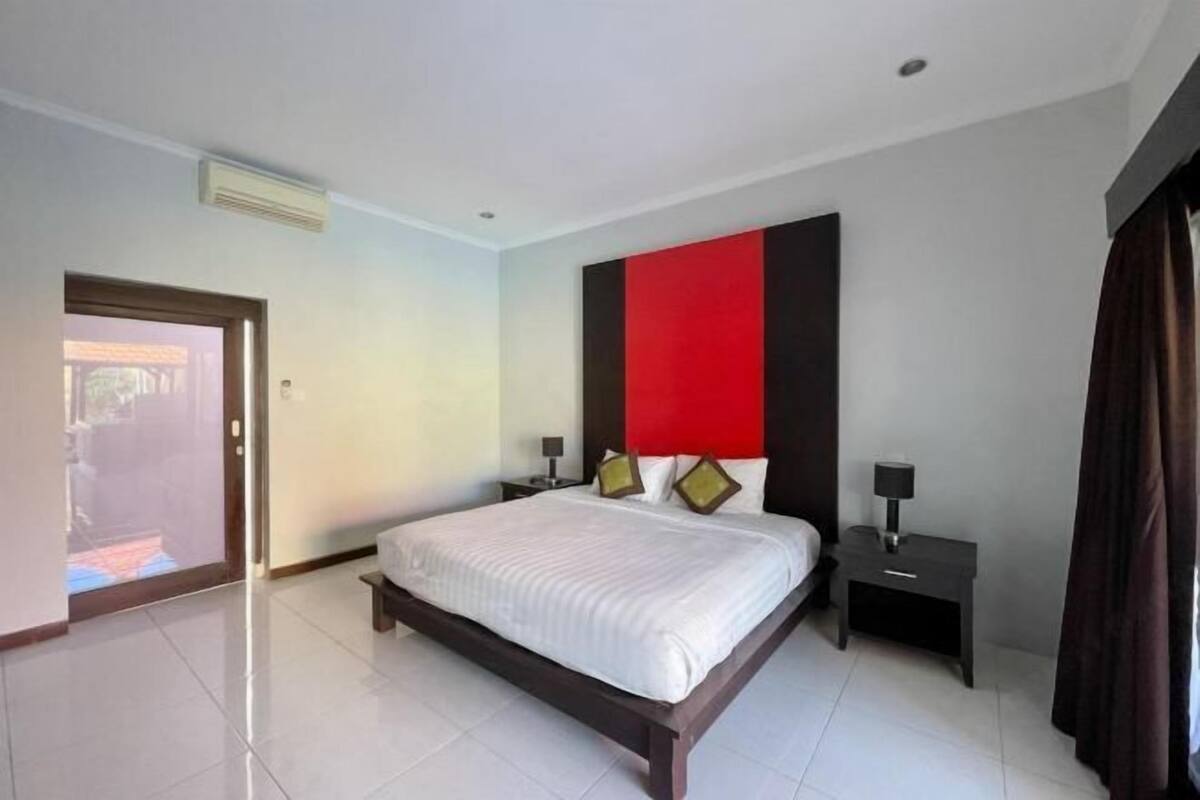 Una habitación de hotel moderna con una cama grande, una pared de acento roja y una mesita de noche pequeña con una lámpara.