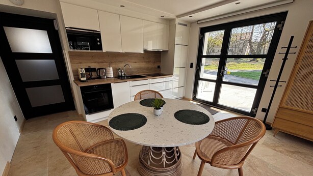 A modern kitchen with white cabinets, a round dining table with woven chairs, and a large window offering a view of the outdoors. - Lodging in a garden near Fraispertuis City Park. (Rambervillers)