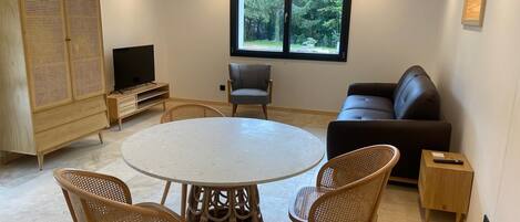 A modern living room with a round table, four wicker chairs, a black leather sofa, a television, and a large window with a view of greenery.