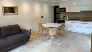 A modern kitchen with white cabinets, a built-in microwave, and a sink. There is a round table with four wicker chairs and a dark-colored sofa in the living area. - Lodging in a garden near Fraispertuis City Park. (Rambervillers)