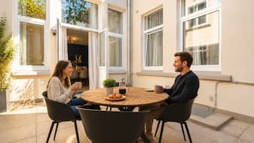 Zwei Personen sitzen an einem runden Tisch mit Kaffeekanne, Gebäck und einer Zimmerpflanze.