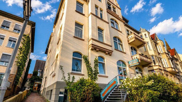 Un edificio de varios pisos con una escalera que lleva a la entrada, rodeado de vegetación y un cielo despejado.