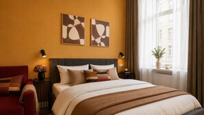 A hotel room with a bed, a red armchair, two wall-mounted artworks, a nightstand with a lamp, and a window with a view of buildings. - Zentrales Studio mit Küche & Waschküche (Bielefeld)