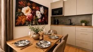 Un comedor con una mesa de madera dispuesta para cuatro personas, un cuadro de flores en la pared, una cocina con armarios y un microondas, y una planta en la encimera.