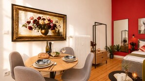 A dining area with a round table, chairs, and a painting of flowers. There is a bed with a red wall and a mirror. - Helles Studio mit Waschküche – Paulusstraße (Bielefeld)
