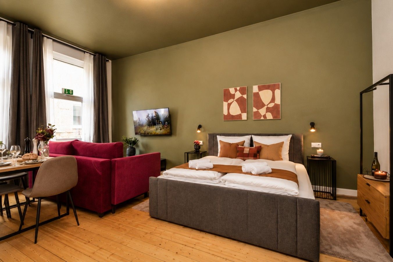 A hotel room with a bed, a desk with a chair, a red sofa, and artwork on the walls.