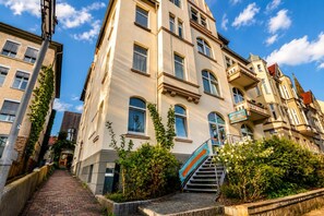 Un edificio de varios pisos con una escalera que conduce a la entrada, rodeado de follaje y un cielo despejado.