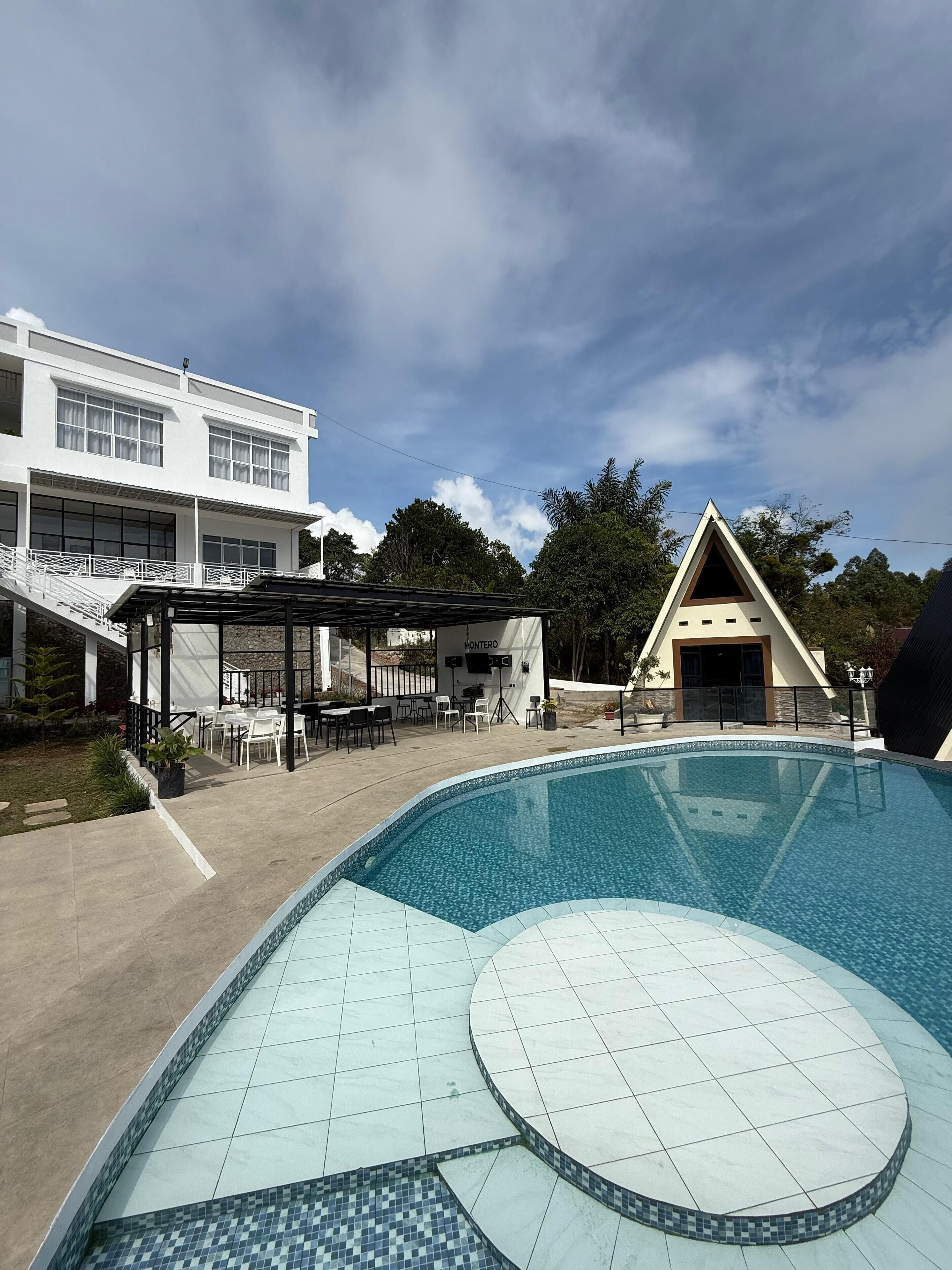 Una piscina con borde de azulejos, un área de estar techada al aire libre y un edificio blanco con ventanas amplias.