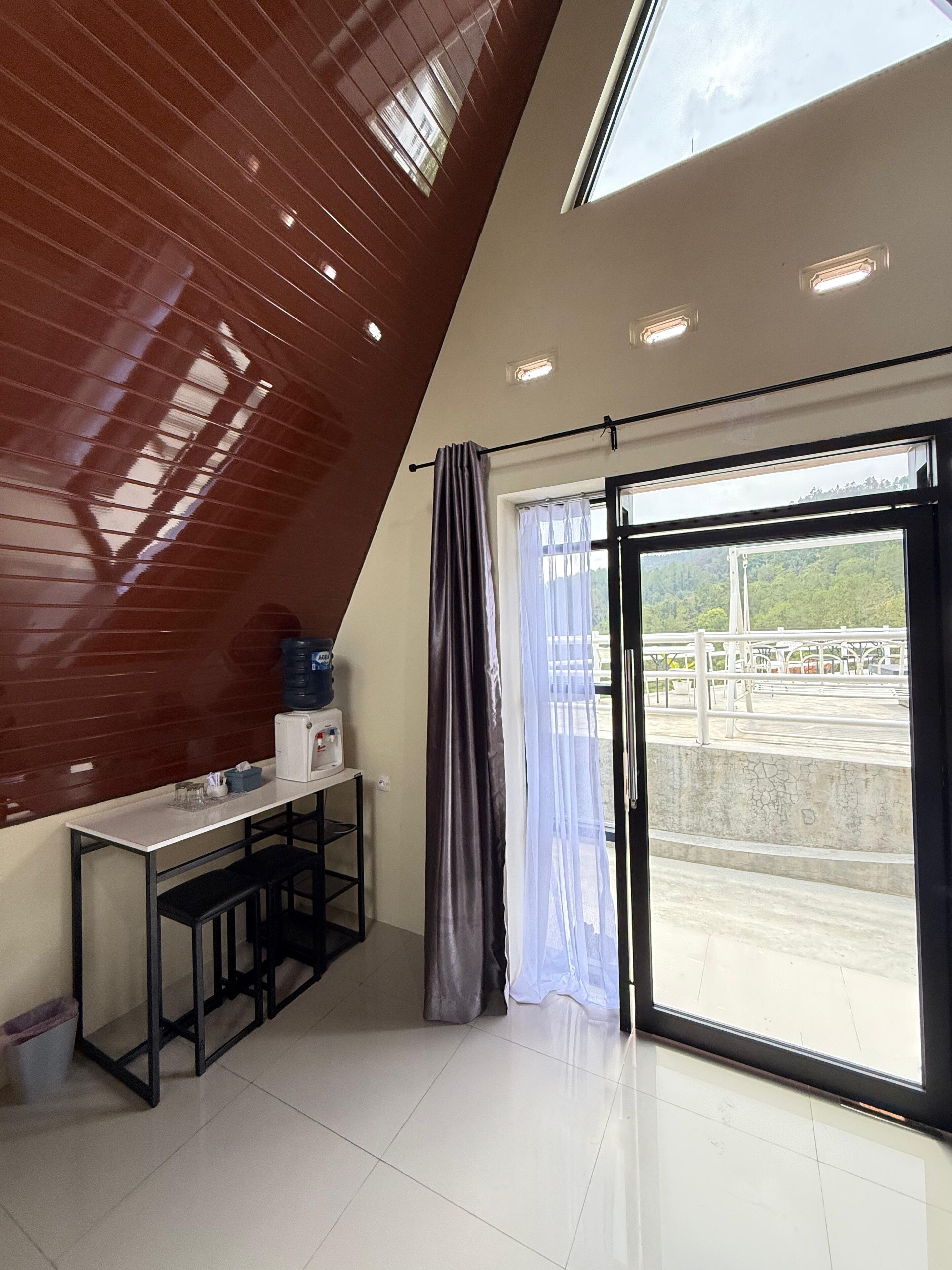 Een kamer met een schuine plafond, een tafel met een waterdispenser, een groot raam met uitzicht op een park en een gordijn.