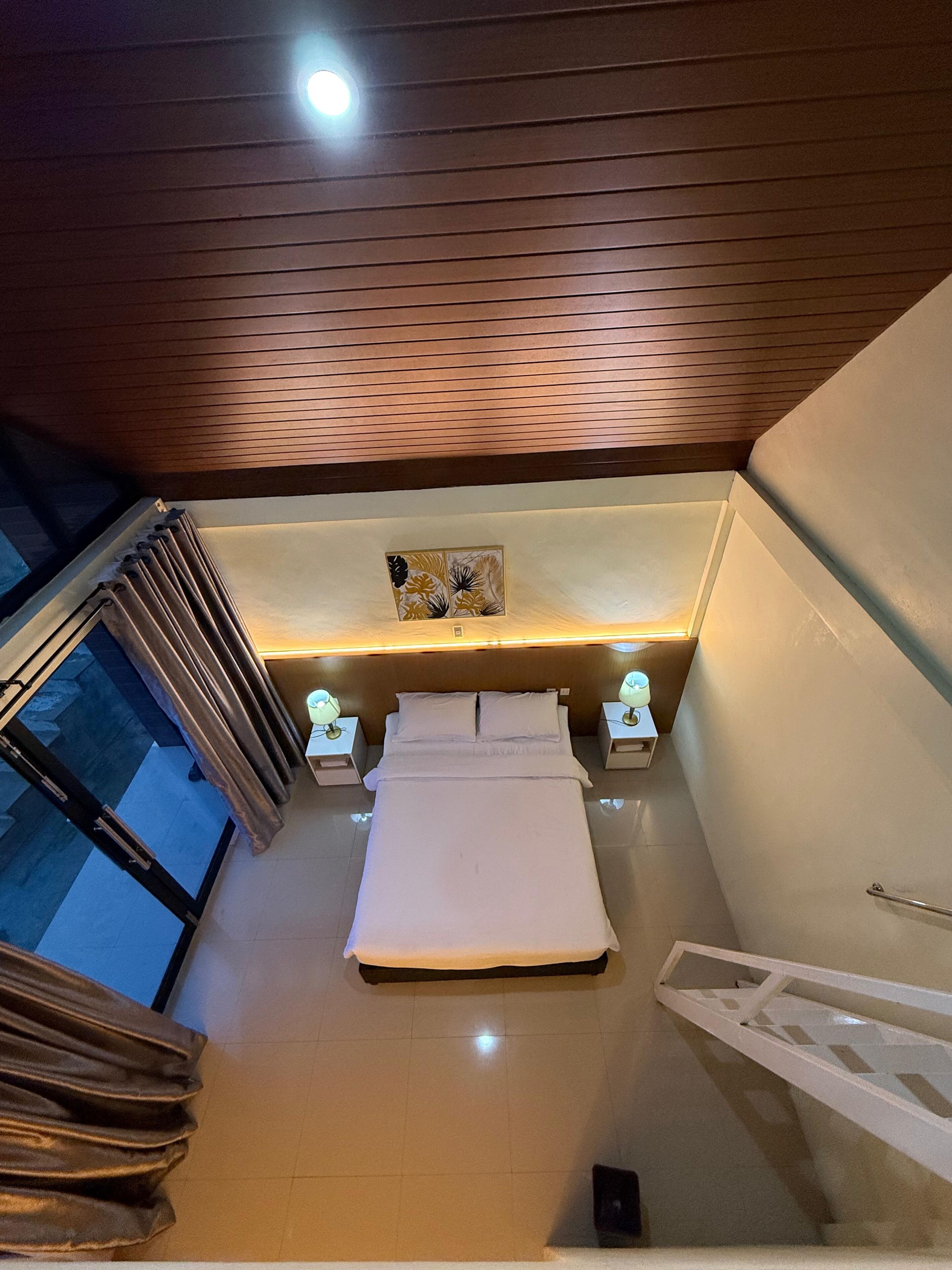 A modern bedroom with a bed centered under a sloped ceiling, flanked by nightstands with lamps, a window with curtains, and a wall-mounted artwork.