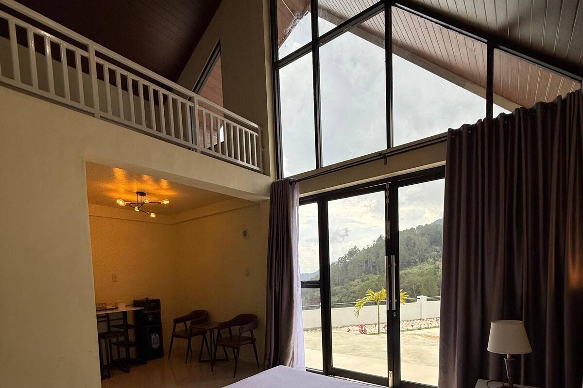 A hotel room with a large bed, a balcony with a view of the outdoors, and a desk area with chairs.