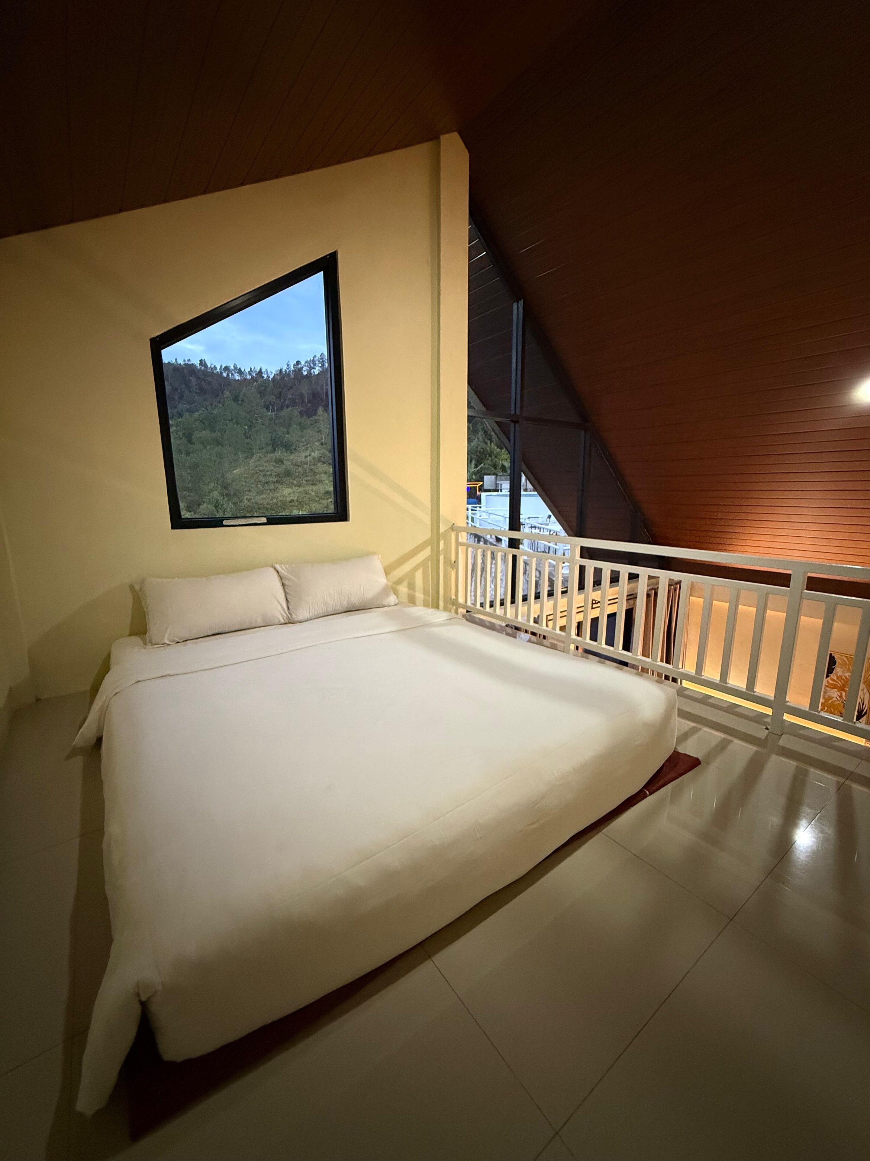 A bedroom with a large bed, a window with a forest view, and a white railing.