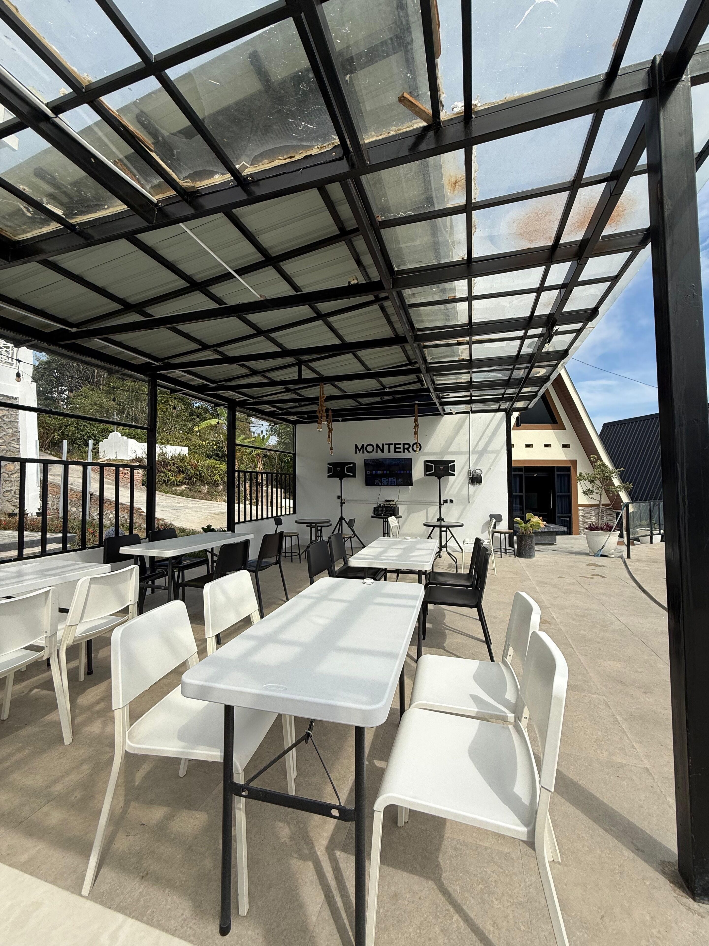 Een buitenpatio met tafels en stoelen, een overdekte glazen structuur en een muur met de merknaam 'MONTERO'.